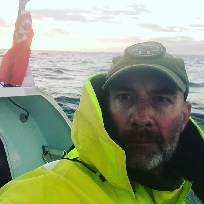 Man in a cap and hi-vis yellow jacket taking a selfie aboard a small boat at sea