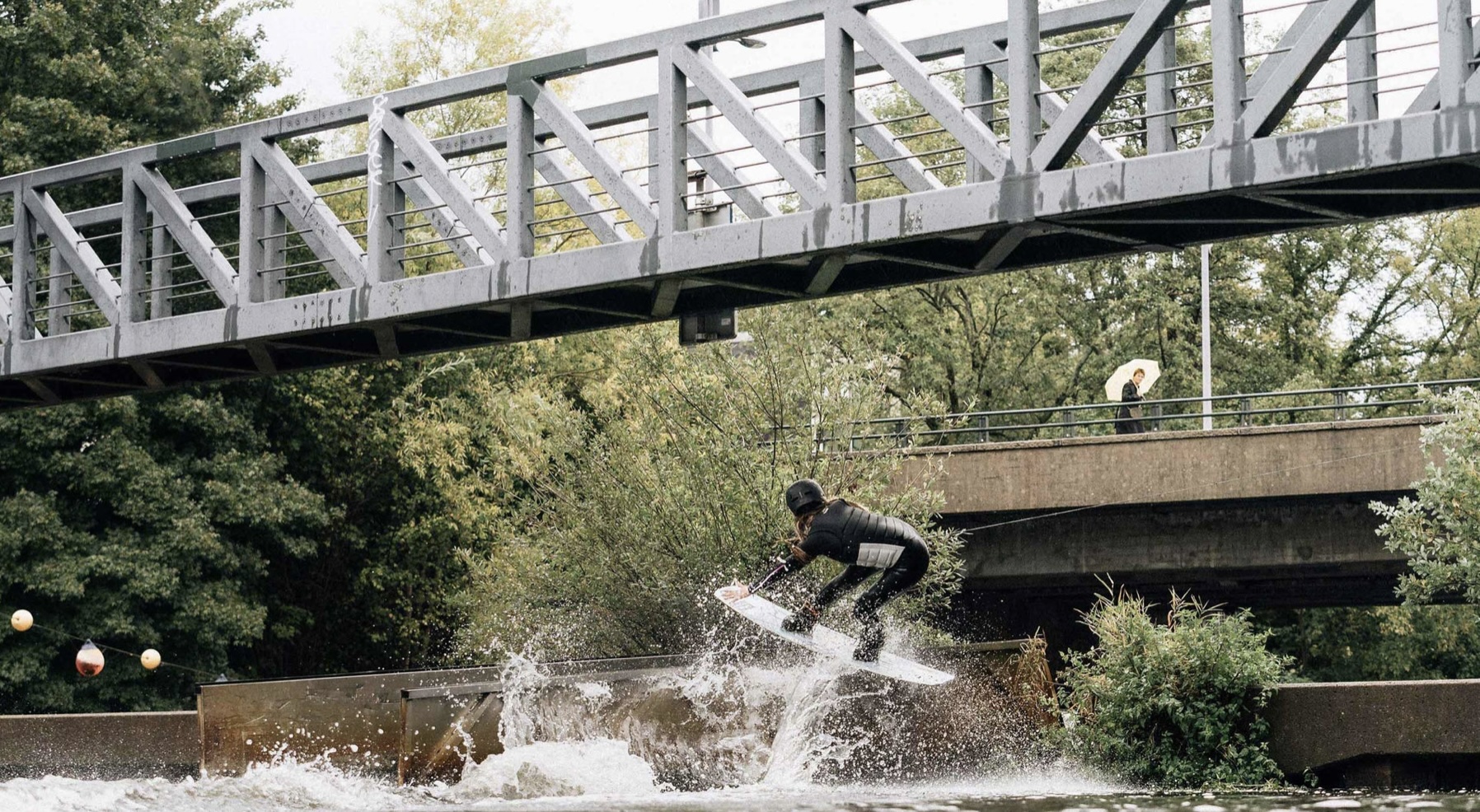 Rider surfing a standing wave on an inland cable park beneath a steel footbridge