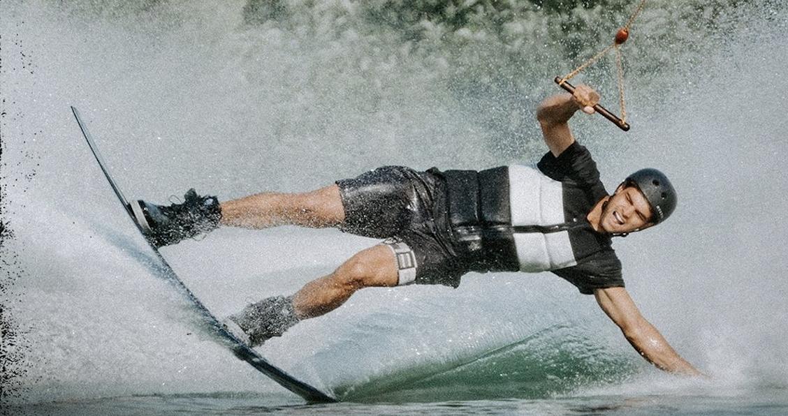 Wakeboarder leaning hard into a turn behind the tow line, wearing a black impact vest and helmet