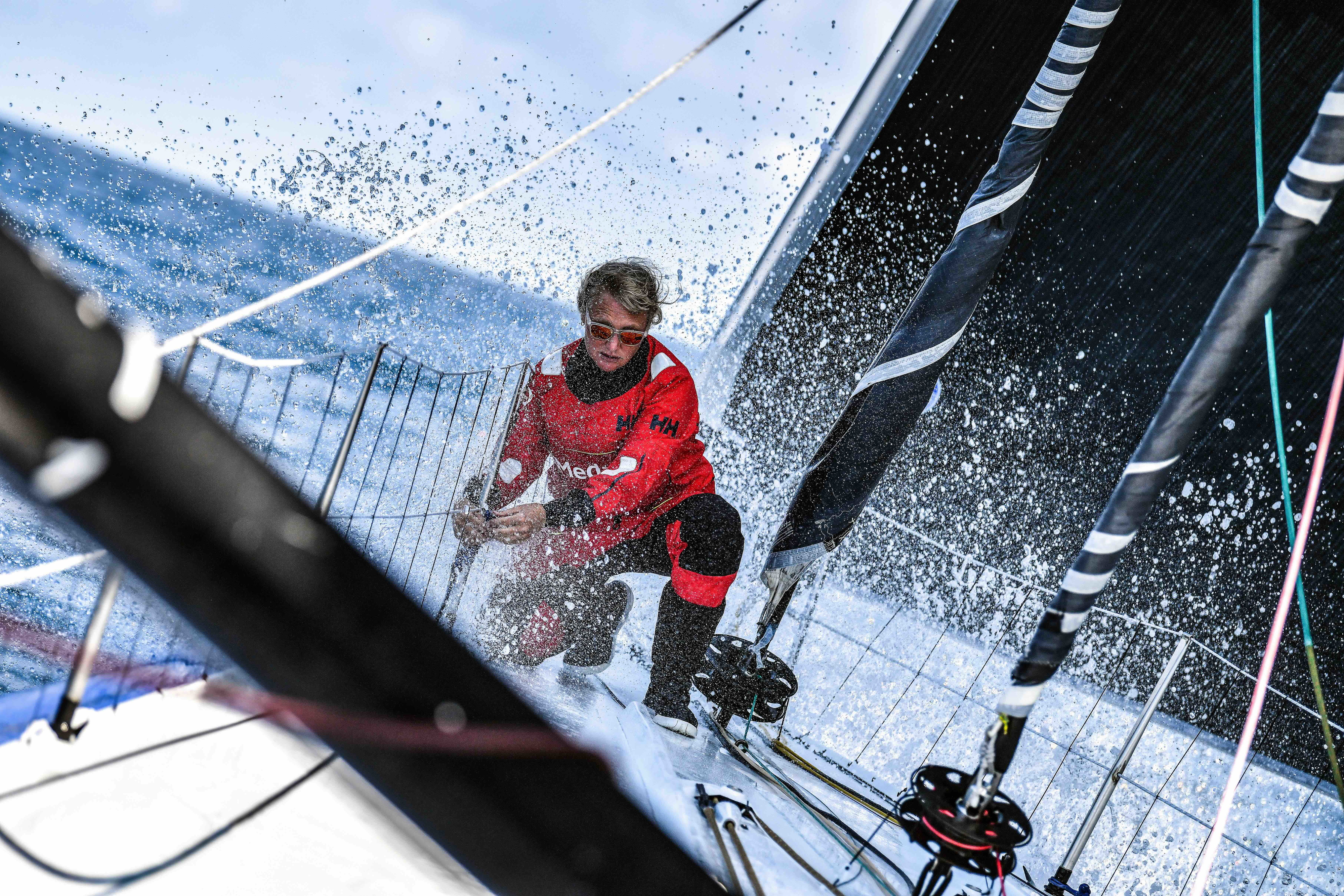 Sailor in a red offshore jacket working on a racing yacht's foredeck amid flying spray
