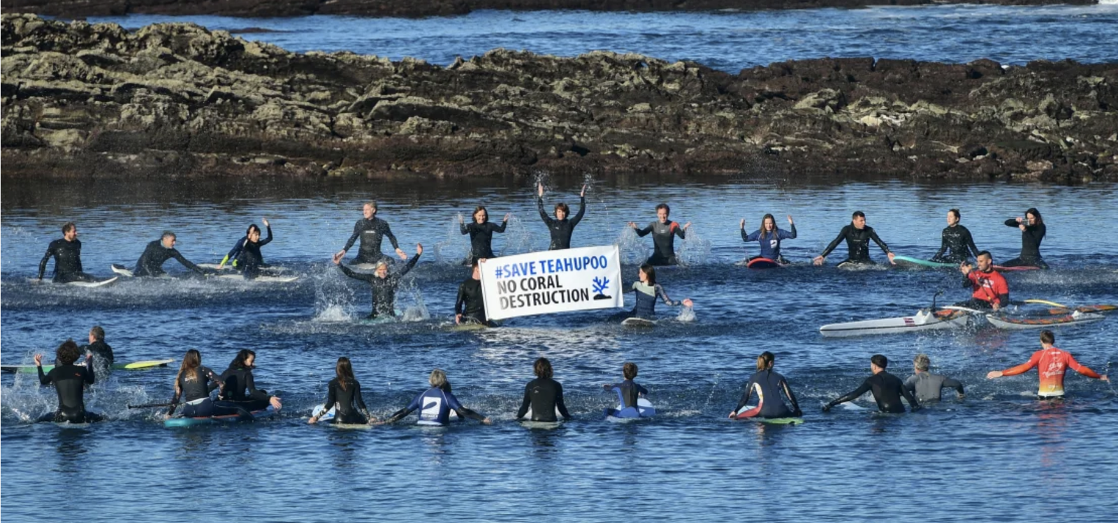 Paddle-out protest with dozens of surfers on boards holding a banner against coral destruction