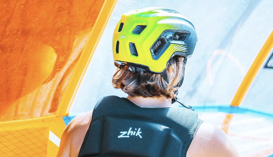 Sailor seen from behind in a yellow helmet and navy Zhik buoyancy aid beside an orange sail
