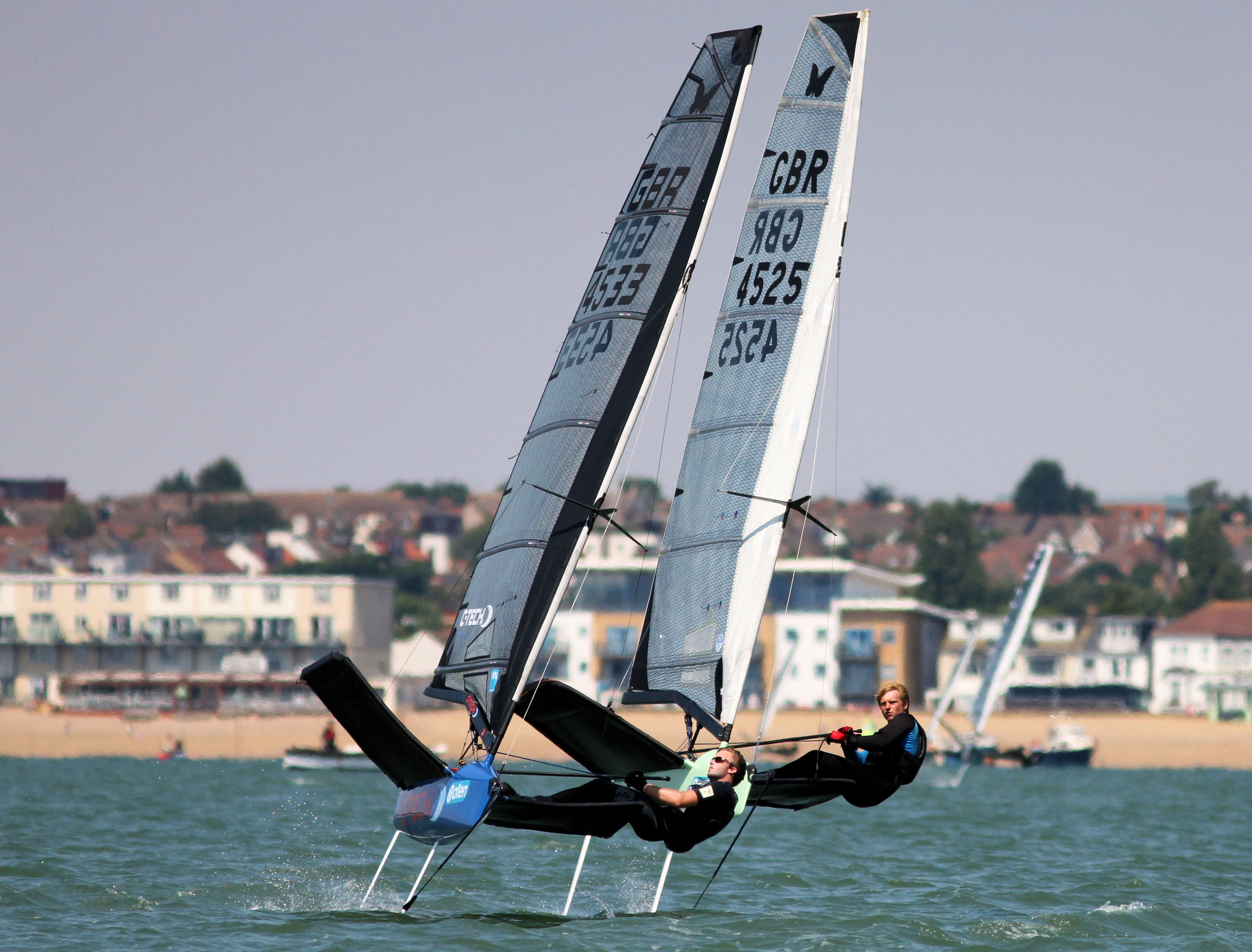 Two foiling Moth dinghies racing side by side in front of a seafront row of houses